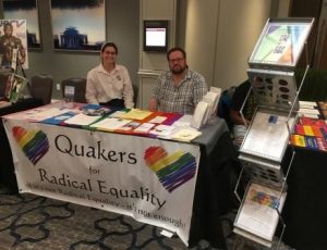 a white woman and white man behind a table with trans, gay, and asexual pride flags, with a sign that says Quakers for Radical Equality
